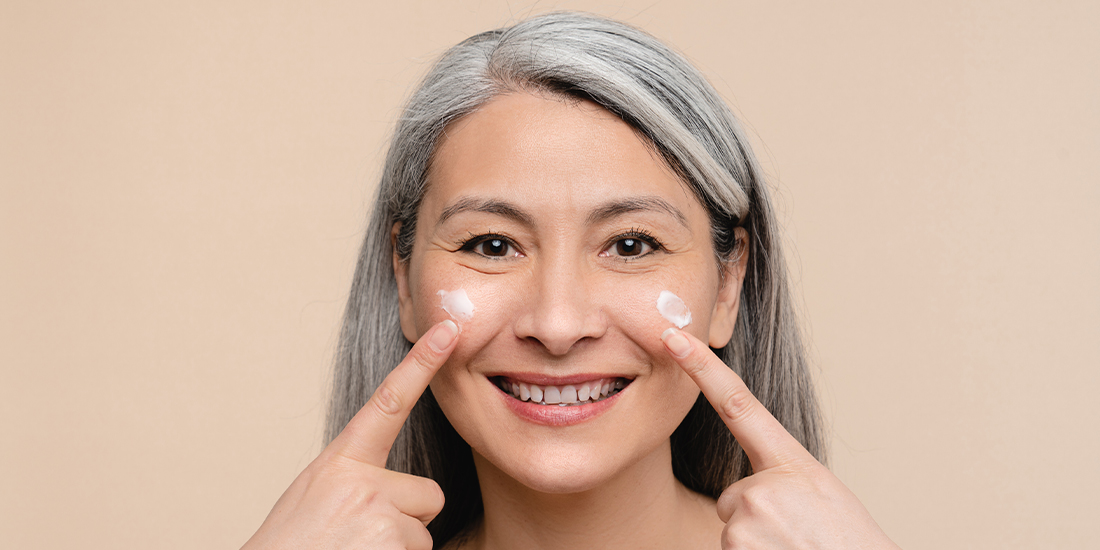 Eine Frau mit langen grauen Haaren lächelt und schmiert sich mit beiden Zeigefingern Creme auf die Wangen. Eine Frau mit langen grauen Haaren lächelt und schmiert sich mit beiden Zeigefingern Creme auf die Wangen.